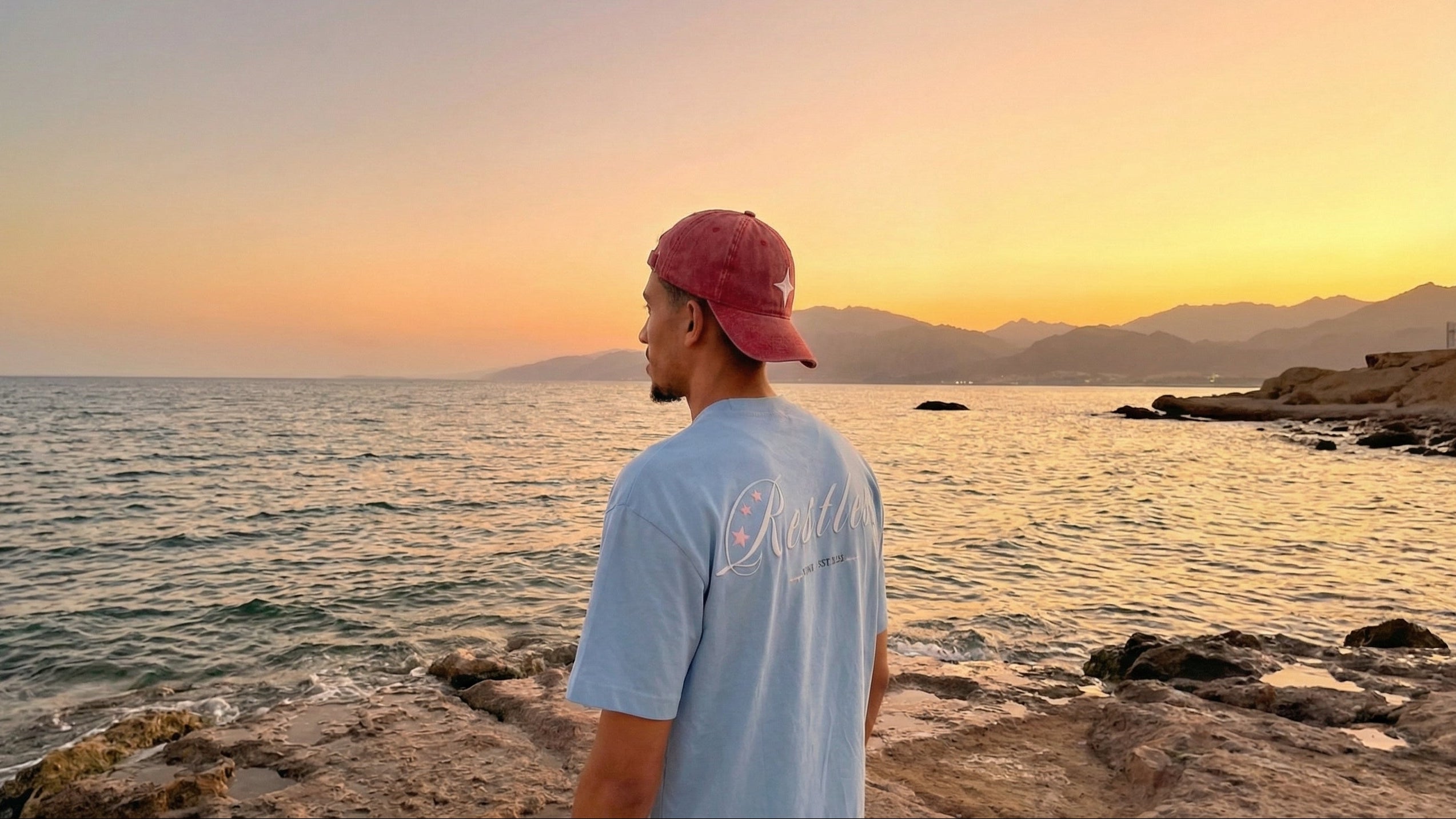 Man wearing a red cap and light blue t-shirt standing on a rocky beach at sunset.